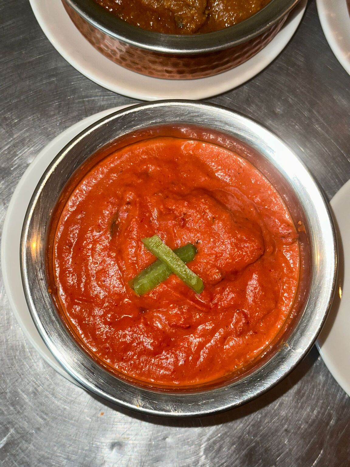 A rich, vibrant red Indian curry, likely Chicken Tikka Masala, served in a 
traditional metal balti bowl at MariGold restaurant in Kilmacolm.